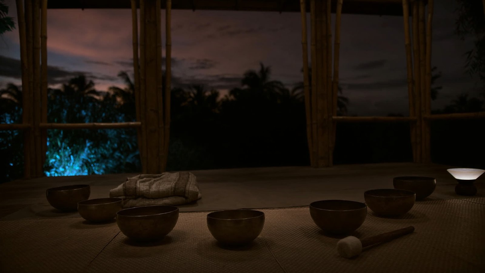 Seven bronze singing bowls arranged in an arc on woven mats in an open pavilion at dusk.